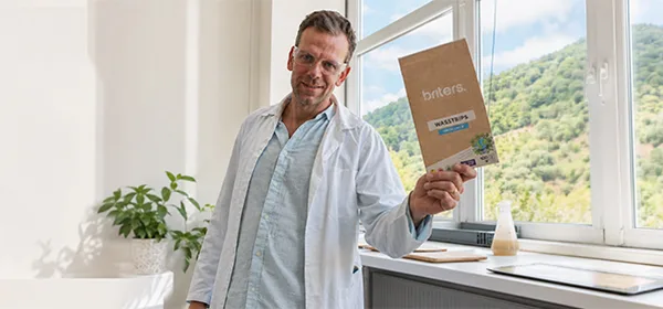 Een man in een laboratoriumjas houdt een verpakking Briters wasstrips vast naast een raam met groene heuvels op de achtergrond