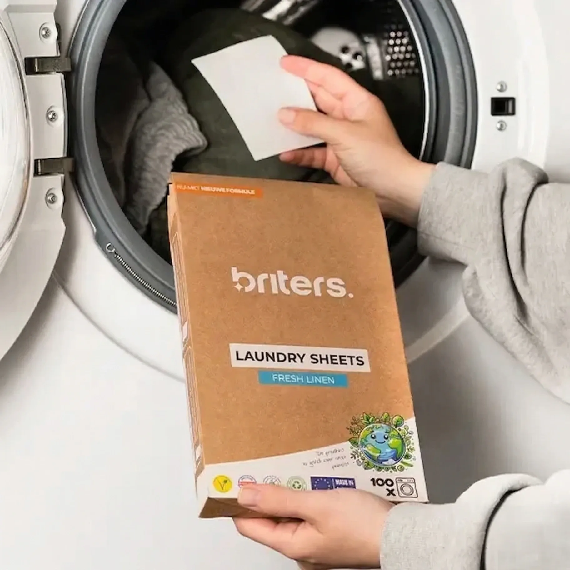 A hand places one laundry sheet in the drum of a washing machine, holding a Briters Fresh Linen pack