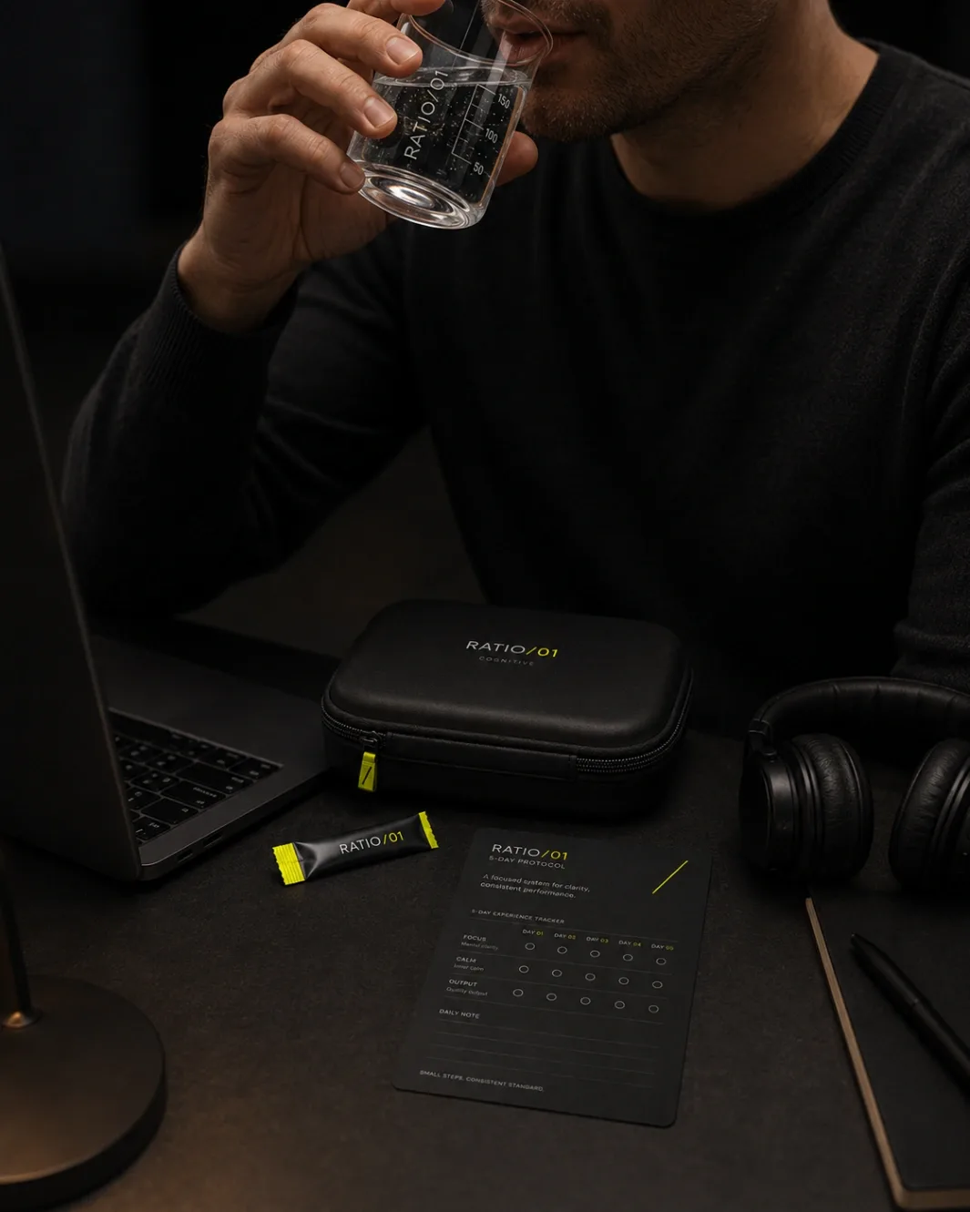 A builder drinking from the marked RATIO/01 lab glass at his desk, closed black case, torn sachet and the 5-Day Protocol card in front of the laptop